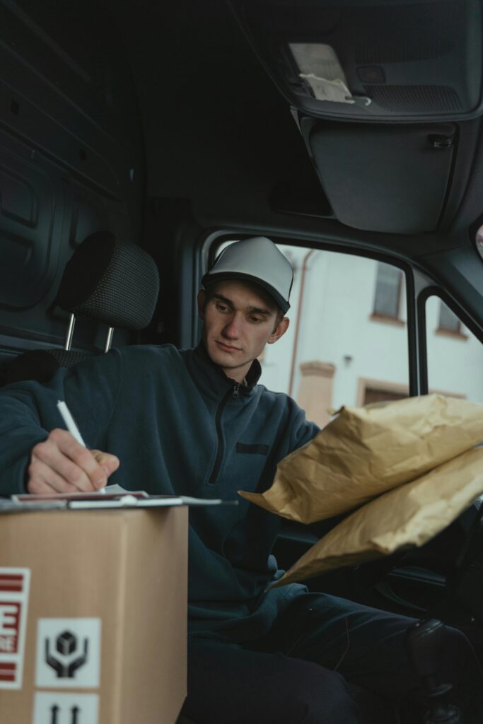 pexels-photo-6169131-6169131 Courier writing on clipboard inside delivery van holding package.