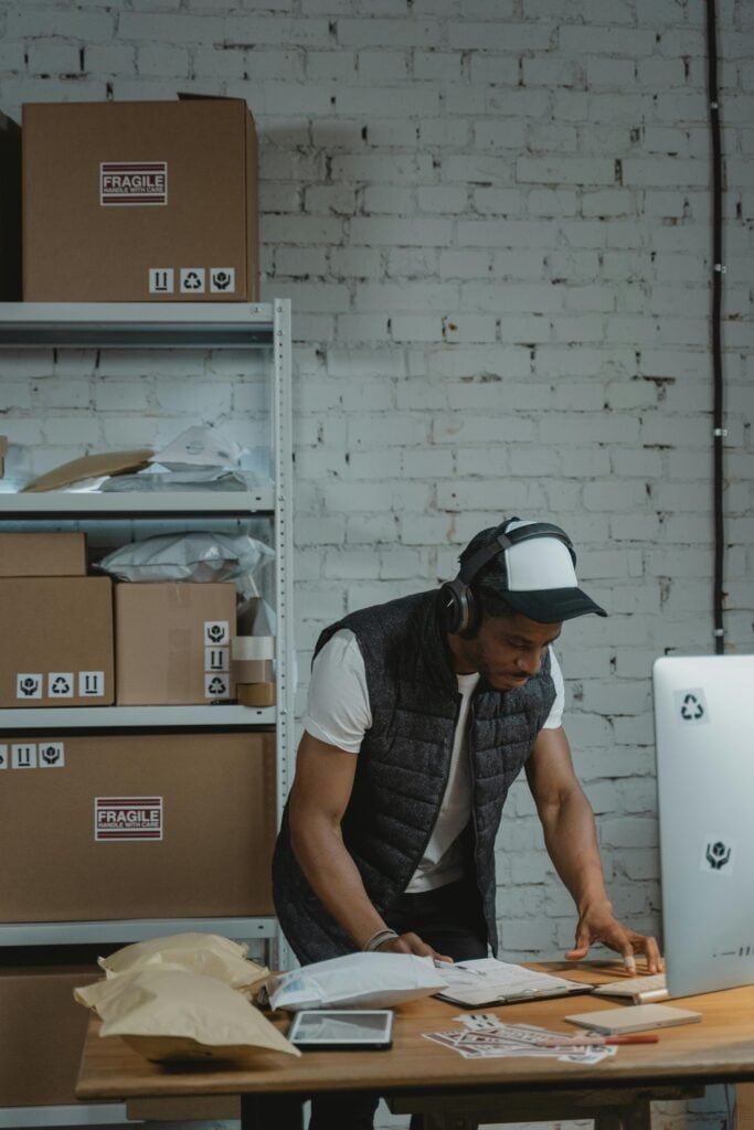 pexels-photo-6170407-6170407 A warehouse worker uses a computer to manage shipments surrounded by parcels and shelves in an industrial setting.