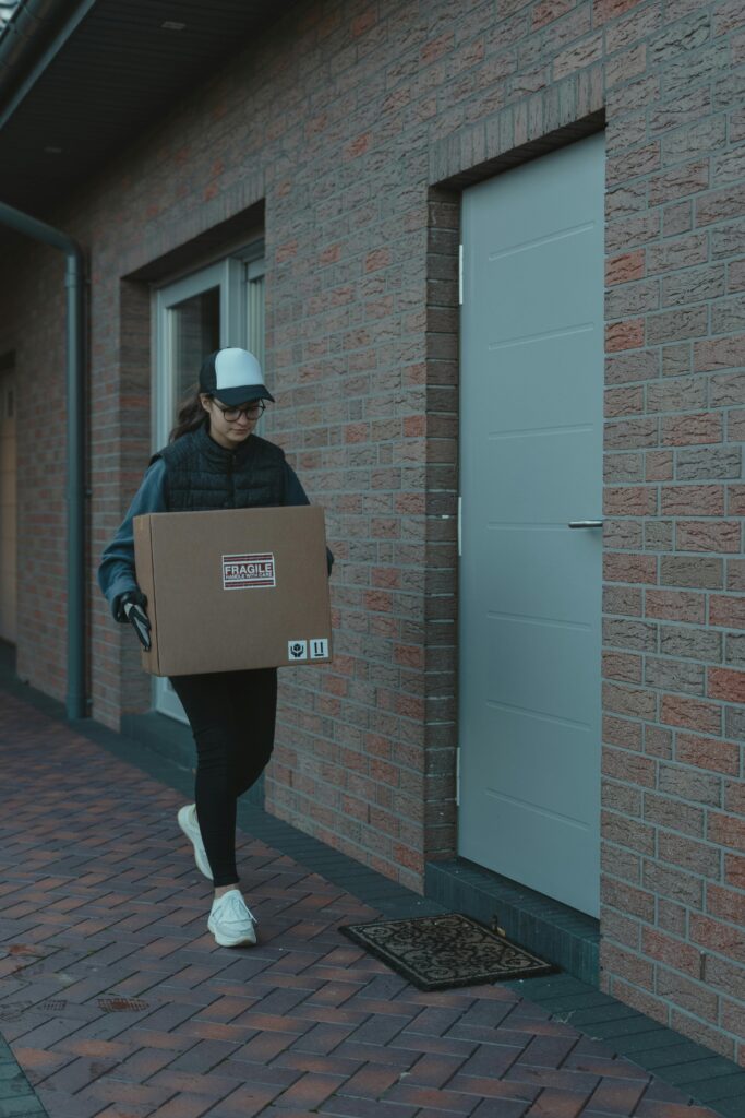 pexels-photo-6170470-6170470 Courier wearing a cap and gloves delivering a cardboard box to a residential doorstep.