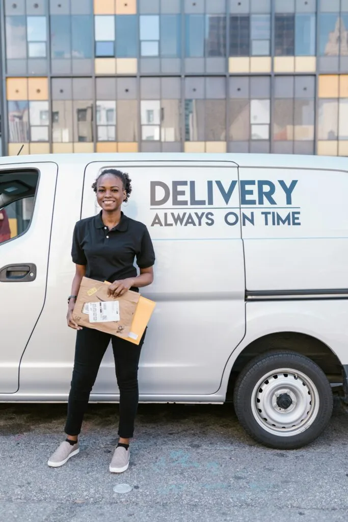 pexels-photo-7362888-7362888 A cheerful woman holding packages in front of a delivery van, symbolizing efficient service.